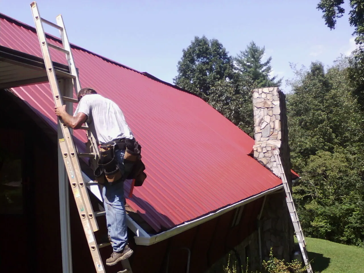 Roofer on a ladder beside a red metal roof and stone chimney — Soffit & Fascia Repair by Roofing Brothers in South Lake Tahoe